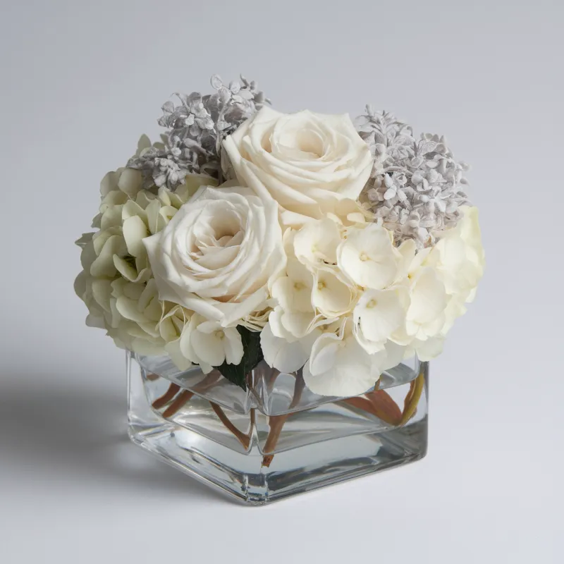An elegant floral arrangement of white hydrangeas, roses, and grey carnations in a clear glass cube vase 
