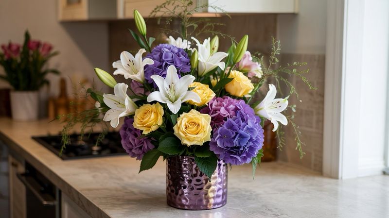 A photograph of a lush floral arrangement, named Meadow Muse, presented in an 8-inch tall, textured metallic purple vase. The bouquet is artfully composed with pristine white lilies, vibrant yellow roses, and voluminous purple hydrangeas accented by wisps of asparagus fern, creating a natural, slightly untamed aesthetic. Positioned on a light yellow kitchen counter, the arrangement is softly illuminated by diffused window light, highlighting the delicate petals and rich colors without harsh shadows. The scene evokes a sense of modern elegance, suggesting a beautiful "Mother’s Day Gift".
