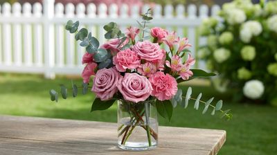 A close-up photograph of the Grad Glow flower arrangement, featuring 5 pink roses, 6 pink carnations, 3 pink alstroemeria, and 4 stems of eucalyptus in a simple glass vase, set on a small table in a sunny backyard. The background includes a white picket fence, blooming flowers, capturing a charming and festive outdoor graduation moment.