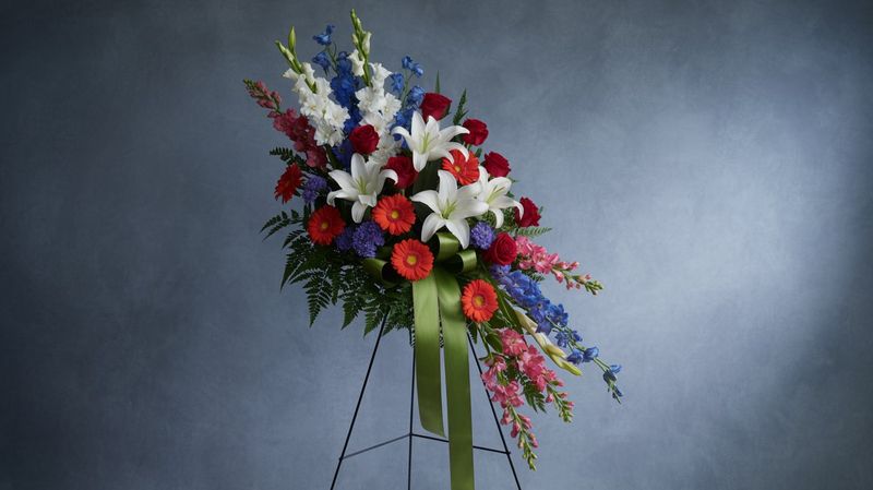 A soft and elegant easel spray with white lilies, red roses, red gerbera daisies, white gladiolus, blue delphinium, pink snapdragons, and greenery like ferns, arranged on a tripod stand with a large green ribbon. The arrangement has a classic, respectful tone with a mix of vibrant and serene colors.  Here is stands against a grey  background in ambient studio lighting.