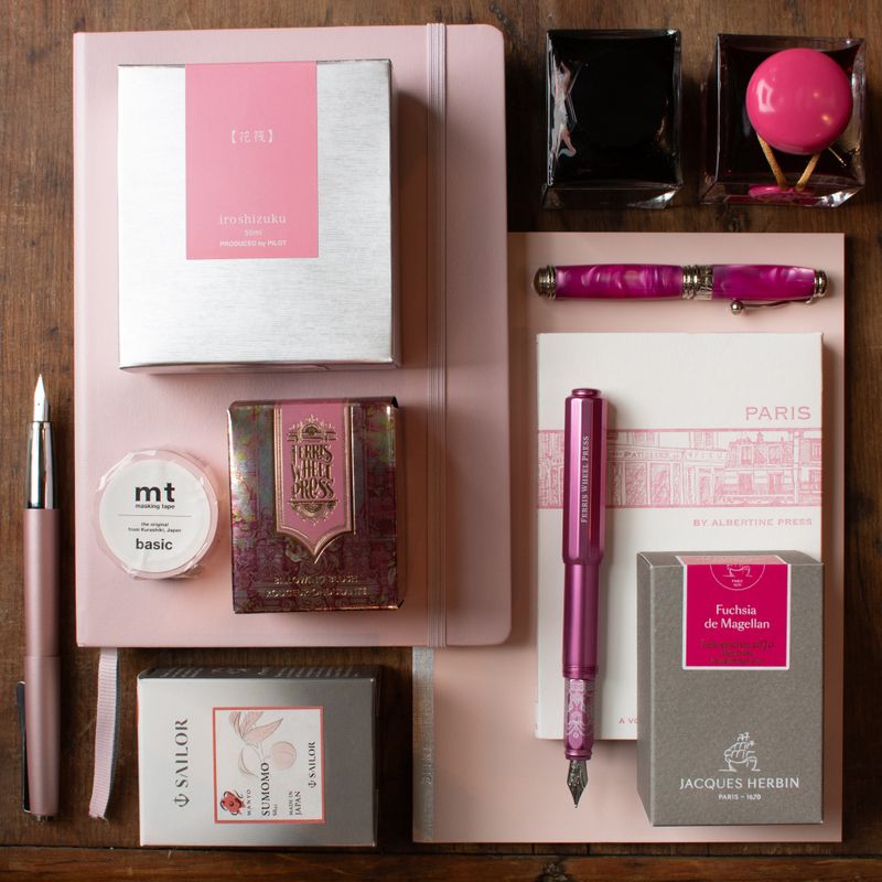 Flat lay of pink fountain pen ink, rose-toned pens, floral-themed notebooks, and writing accessories arranged on a wooden background