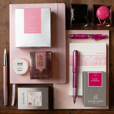Flat lay of pink fountain pen ink, rose-toned pens, floral-themed notebooks, and writing accessories arranged on a wooden background