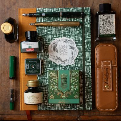 Flat lay of green fountain pen ink, brass-accented pen, sketchbook, and nature-themed accessories styled with leather pen accessories on a wooden background
