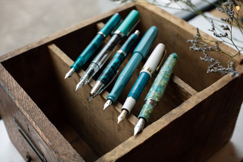 A row of teal and turquoise-toned fountain pens neatly arranged on a wooden box, with sprigs of dried flowers.