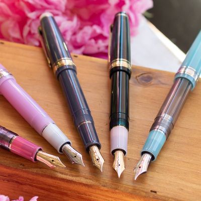 Close-up of colorful fountain pens with gold nibs displayed on a wooden surface, with pink flowers in the background.