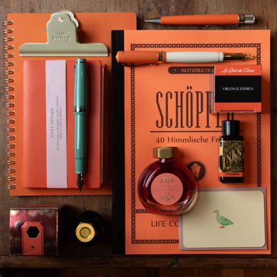 Flat lay of orange fountain pen ink, brass pen, retro-style notebook, and vintage desk accessories on a wooden background.