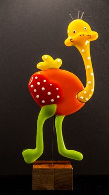 Glasfigur, struds, orange krop, gult hoved, rød vinge , gul hale, grønne ben, går mod højre på træfod