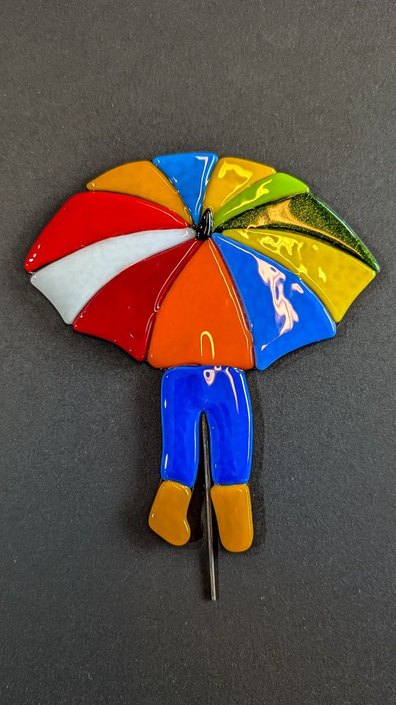 Glasfigur, Paraply dreng i mange farver med blå bukser og orange sko, på træfod Glasfigur, Paraply dreng i mange farver med blå bukser og orange sko, på træfod