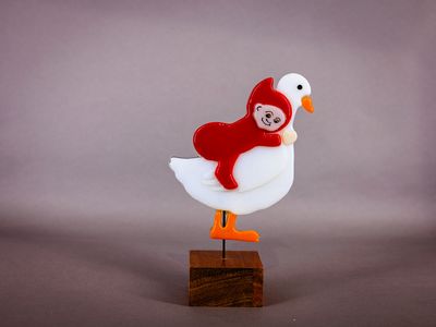 Glasfigur, Nisse riddende på ryggen af gås, vender mod højre, på træfod Håndlavet glasfigur med rød nisse, der ligger på ryggen af en hvid gås med orange næb og fødder, på træfod.