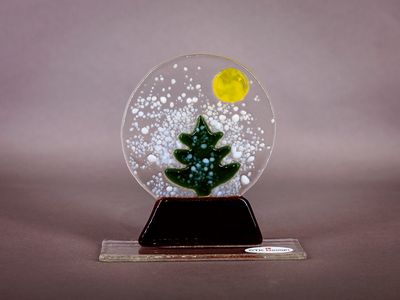 Glasfigur, snekugle, kane med gaver, på glasfod Rundt glasmotiv med grønt grantræ, hvid sne og gul måne på mørk fod.