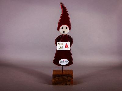 Slank nissepige i mørkerødt glas med lang spids hue og et lille God Jul-skilt, monteret på træfod.