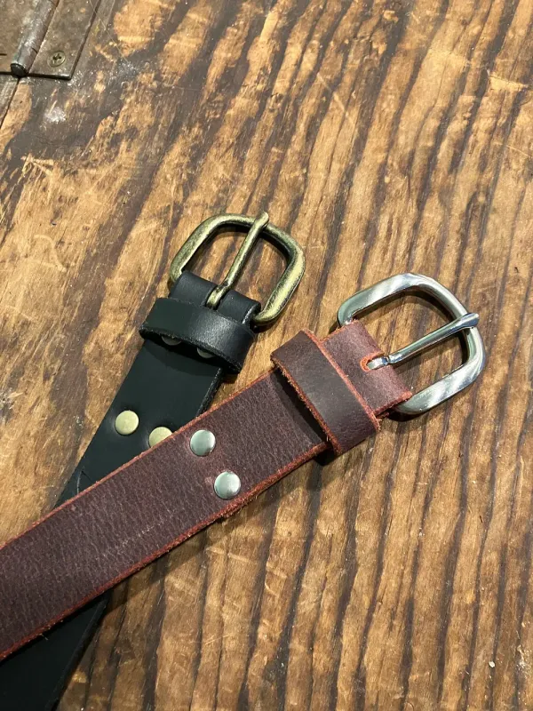 Two 1 inch belts displayed on rustic wood - one black leather with antique brass hardware, the other oxblood with silver