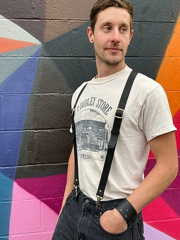 A masculine person standing in front of a street mural wearing suspenders in a casual way