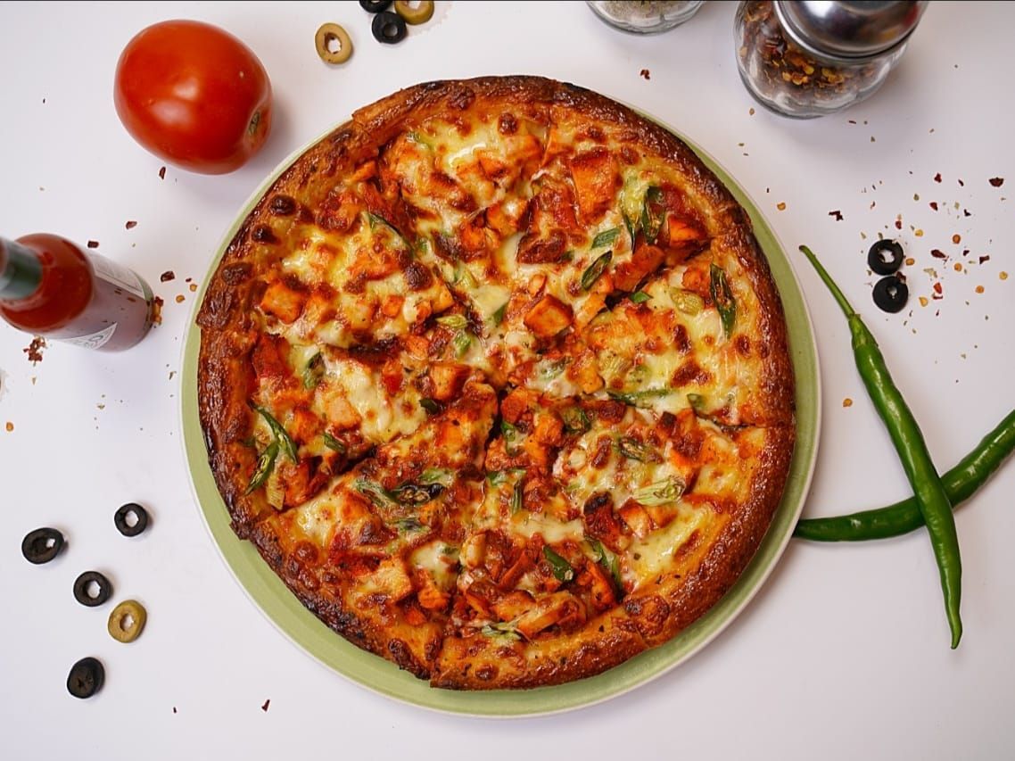 A delicious Chicken Tandoori Pizza from Pizza Corner, featuring a crispy crust loaded with juicy tandoori-spiced chicken, melted cheese, red onions, and green bell peppers. The vibrant toppings are garnished with fresh cilantro, with the cheese bubbling golden around the edges. Served hot on a rustic wooden board, ready to enjoy.