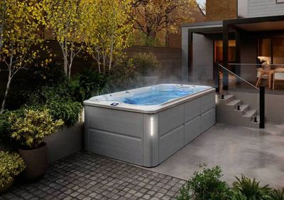 Spa de nage jacuzzi intérieur blanc et extérieur gris  sur terrasse grise avec arbre et verrendant 