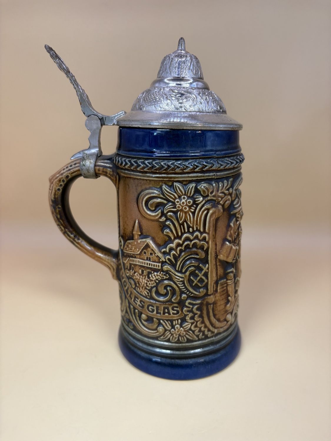 Stein. Germany. Brown w/ Dark Blue Outline. Two Men and a Woman Dancing w/ Beer. Floral Decoration and Houses on Sides w/ Text &quot;Ein Volles Glas&quot; &quot;Des Gastes Spab.&quot; Pewter Lid. 7.5&quot;H