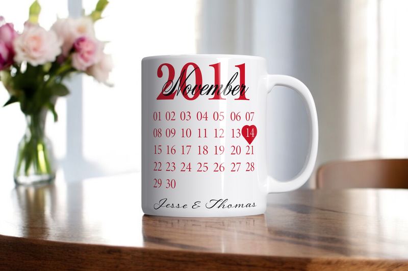 Witte kalender-mok met gepersonaliseerde namen en een belangrijke datum gemarkeerd met een rood hartje, geplaatst op een houten tafel met bloemen op de achtergrond.