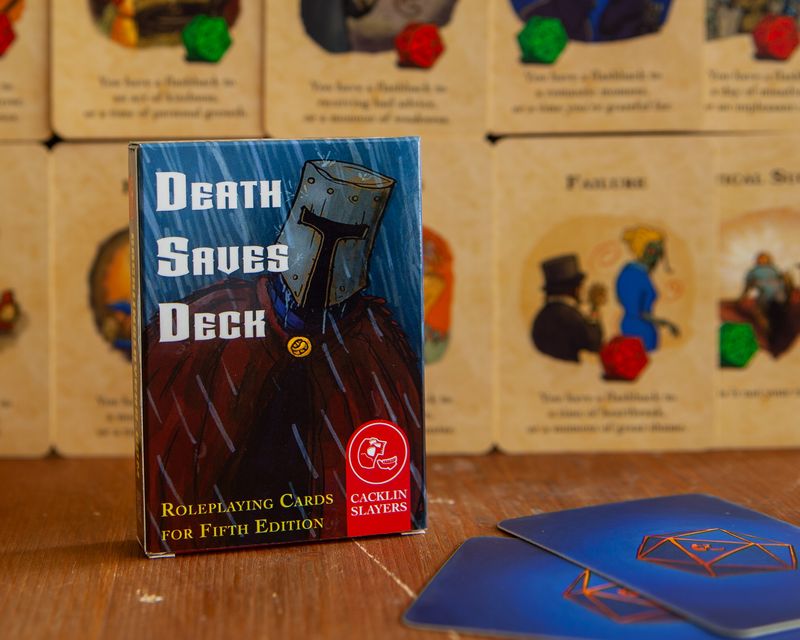 The packaging of Cacklin Slayers' Death Saves Deck features a gloomy knight walking through the rain. The text at the bottom reads "Roleplaying Cards for Fifth Edition."