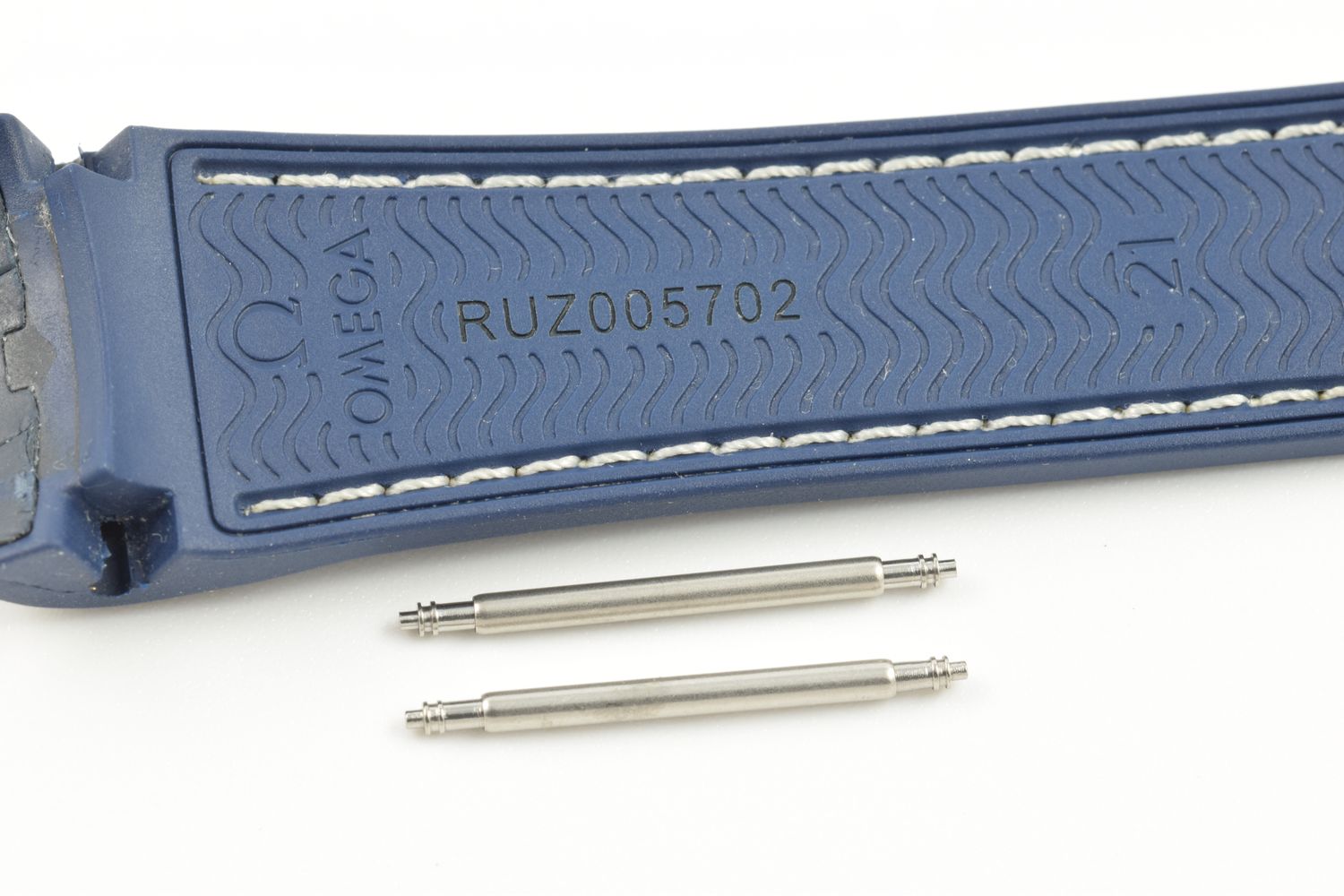 Two stainless steel spring bars placed below a blue Omega Planet Ocean rubber strap stamped RUZ005702 on the underside, shown on a white background