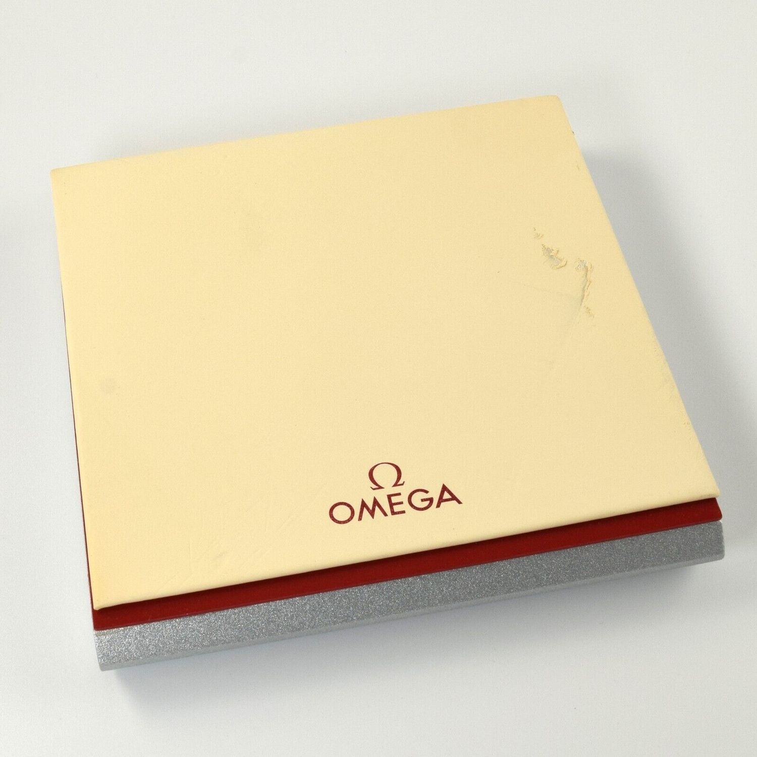 Authentic Omega watch display tray with cream top, red trim and silver base showing a tear on the top surface and red Omega logo printed on the cream section