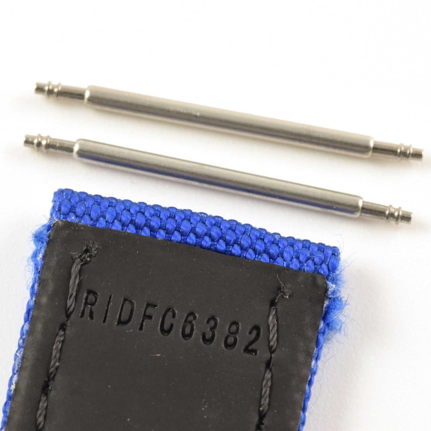 Two spring bars positioned above the end of a blue textile watch strap with black underside stamped RIDFC6382