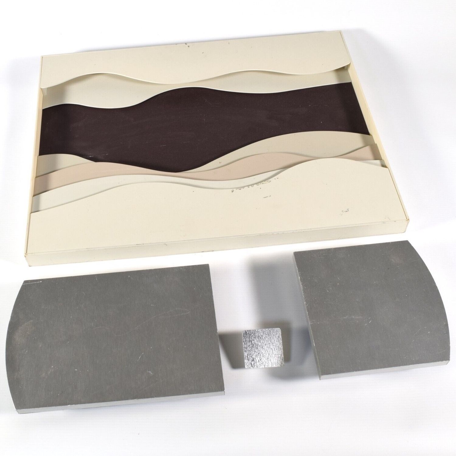 Watch shop display tray set including a cream and brown wave-style tray with scuff marks, two brushed steel curved podiums, and a small metal display cube on a white background