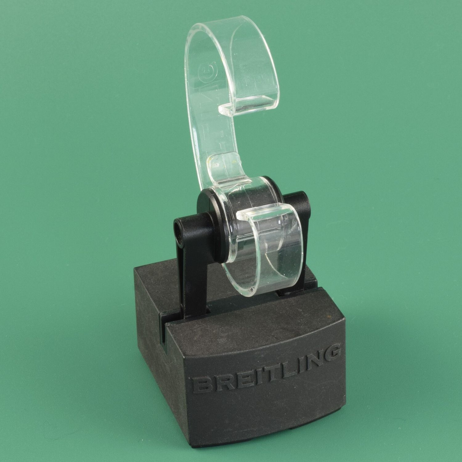 Genuine Breitling watch display stand with a small 3cm high black base and a clear curved plastic watch holder, shown against a green background with BREITLING embossed on the base