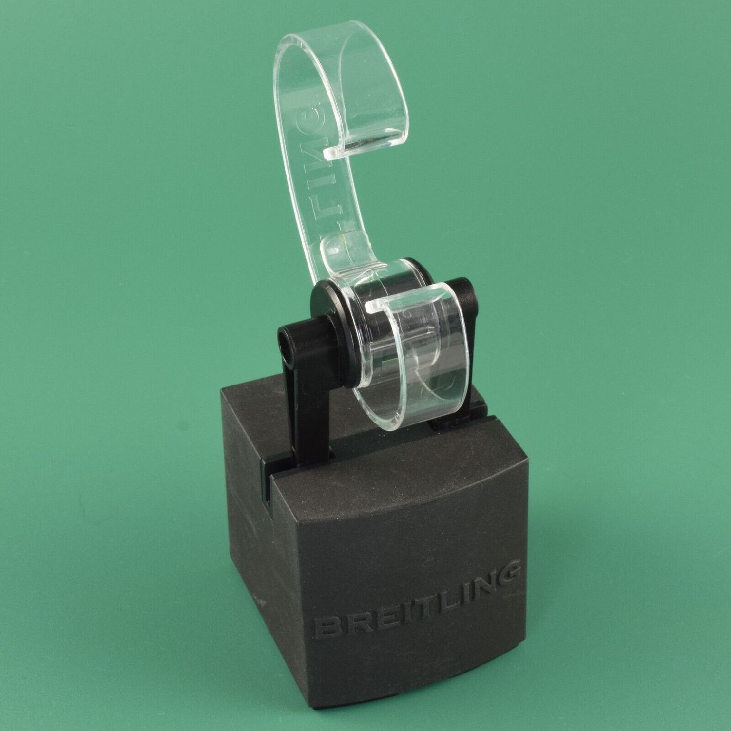 Authentic Breitling watch display stand with a medium 5cm tall black base and clear curved plastic watch holder embossed with the Breitling name shown on a green background