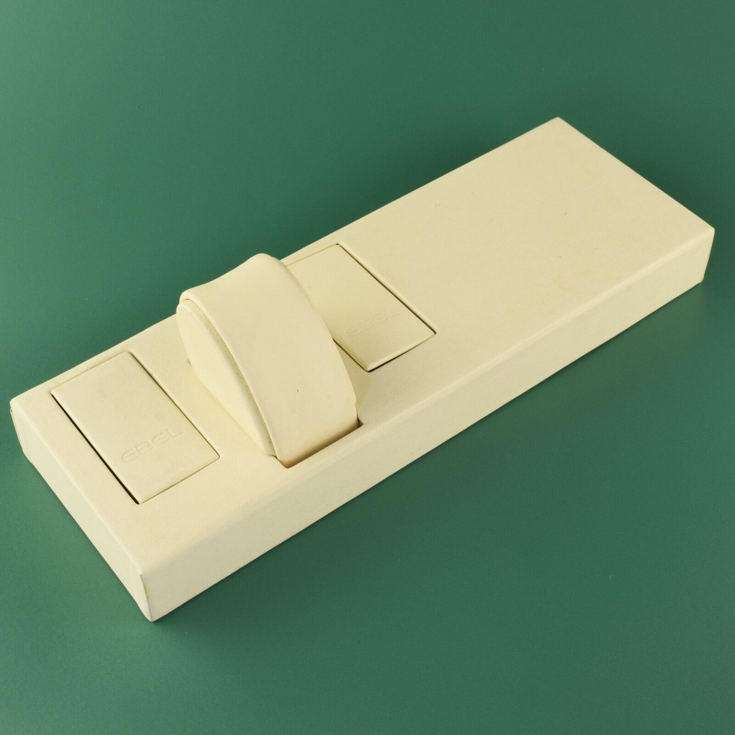 EBEL cream leather display tray with one round watch pillow and two EBEL-logo socket covers on green background