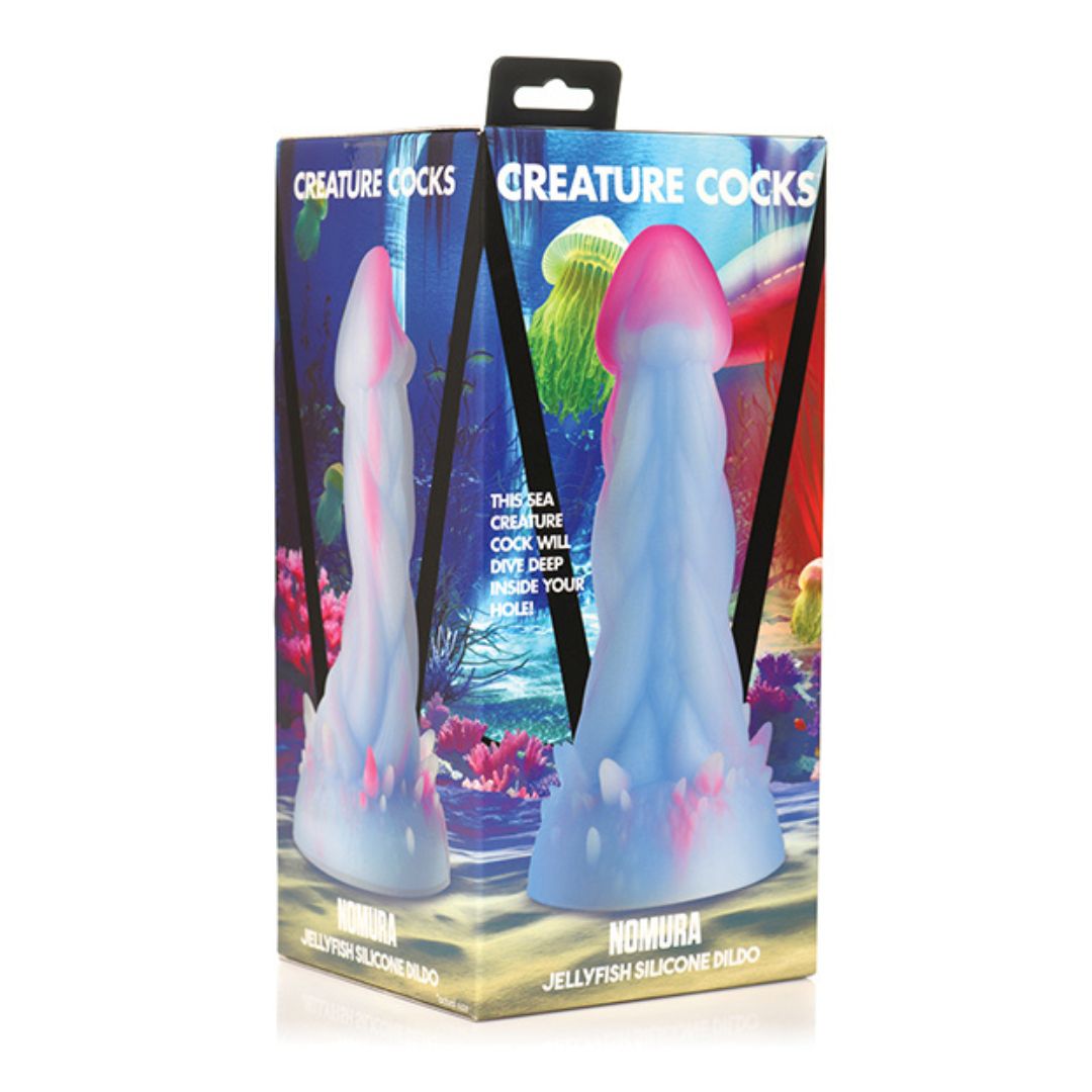The Creature Cocks Nomura Jellyfish Dildo in its box, depicted in front of an ocean of colorful jellyfish. The silicone toy resembles a jellyfish itself, layered with sleek, trailing tentacles with a prominent phallic head and a thick, spiky base. It is primarily a deep ocean blue, with purple marking the tip and scattered throughout. 