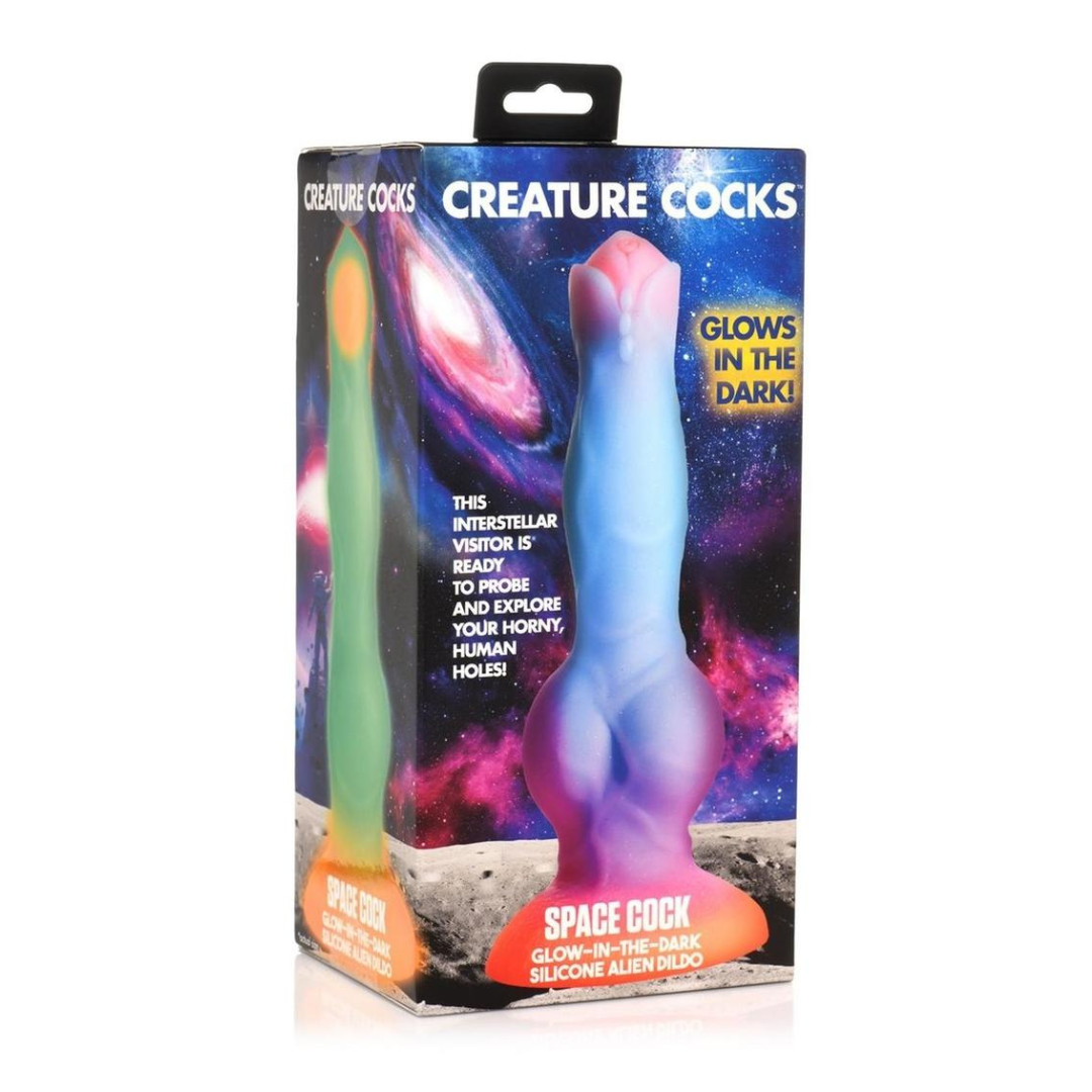 The creature cocks space cock glow in the dark dildo in its box, the toy depicted in front of a colorful galaxy background. The dido itself is poured in a gradient from orange at the base to purple, to blue, to pink at the tip. It has a fairly flat head and a knot near its base.