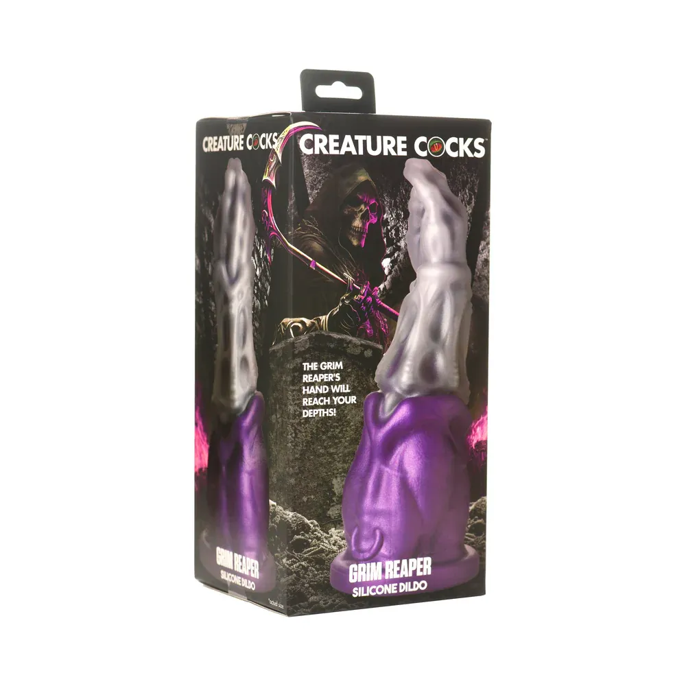 The Creature Cocks Grim Reaper Silicone Dildo shown in its box, depicted in front of an illustration of a grim reaper. The toy is made to resemble the hand and arm of the grim positioned for fisting, poured in glittering silver to represent bone and deep purple lower down to resemble his cloak. The toy bears a prominent skeletal texture, with a tapered tip for easy insertion and an incredibly girthy base. 