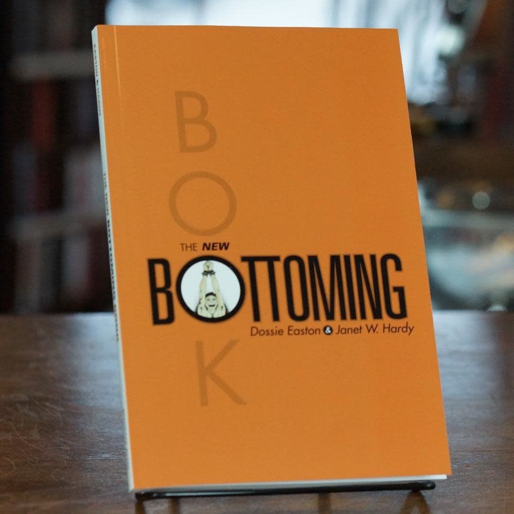 The New Bottoming book - a thin, burnt-orange volume with black + gray text and an image of a happy bottom with bound hands