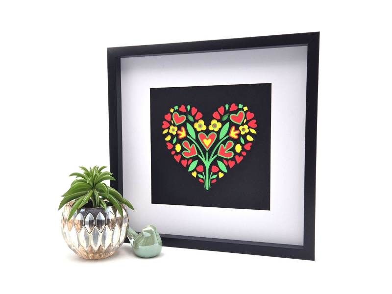 Vierkant schilderij in zwarte lijst met motief van een hart en bloemen op zwarte achtergrond.