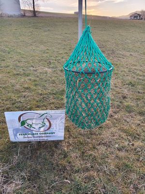 Grünes Heunetz mit Ring, 5kg, für Pferde, hängt an einem Pfosten auf einer Wiese mit einem Schild v…