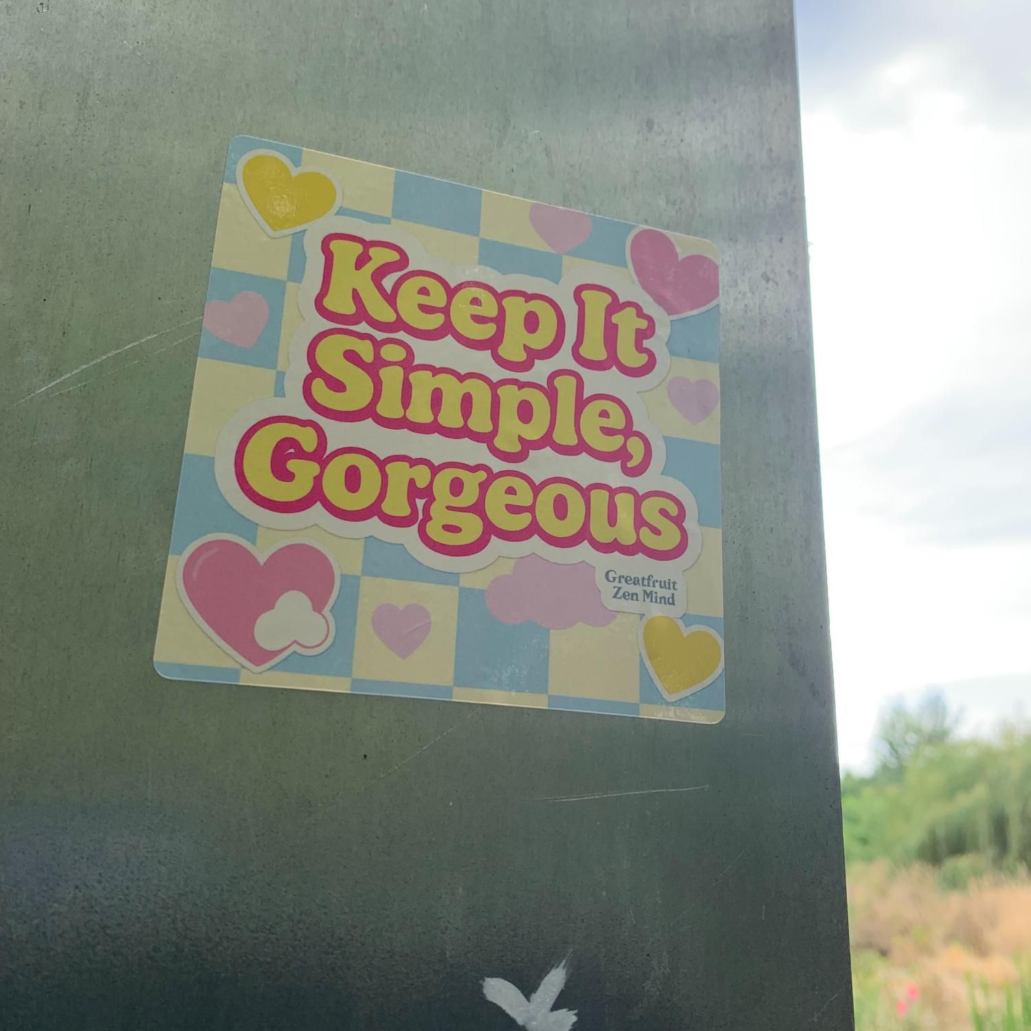 Large Sticker—Keep It Simple Gorgeous. Large ‘Keep It Simple, Gorgeous’ sticker with pastel checkered background and bold yellow text on a sign in nature landscape.