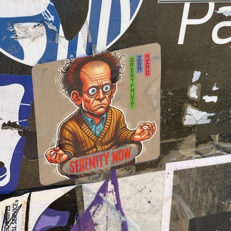 Sticker category image features stressed out meditating man 'Serenity now' sticker set upon a wall of graffiti.