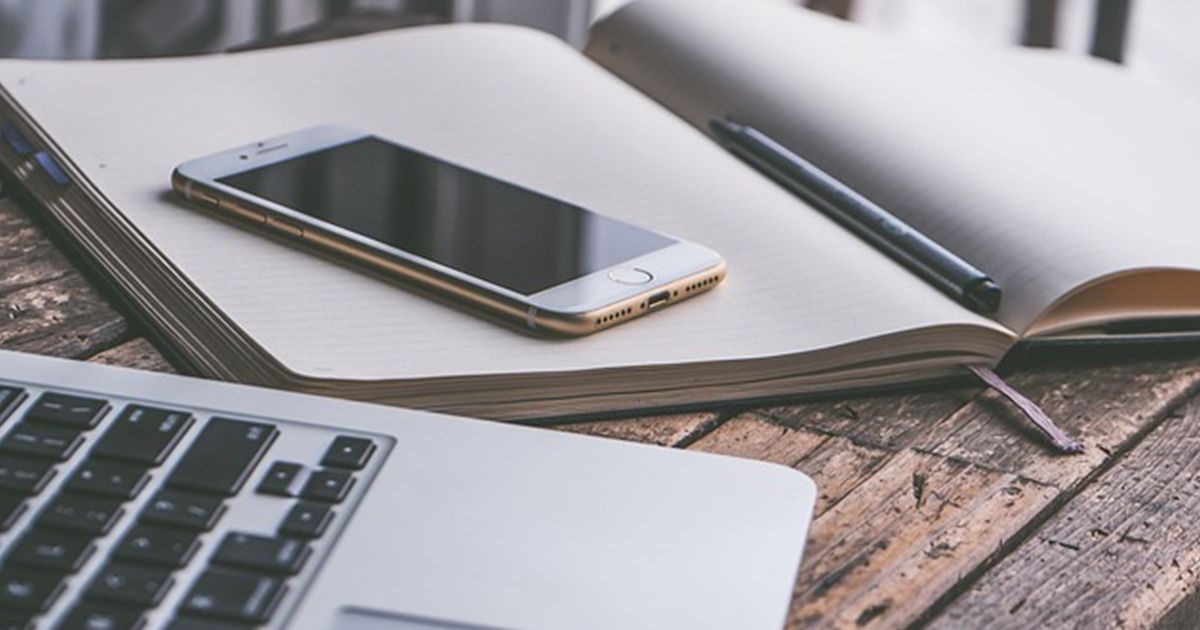 Offenes Notizbuch mit Smartphone, Stift und Teil eines Laptops auf einem rustikalen Holztisch.
