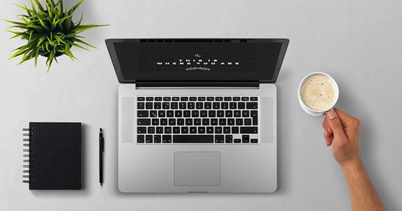 Arbeitsplatz mit aufgeklapptem Laptop, Kaffeetasse in einer Hand, Notizbuch, Stift und Pflanze auf hellem Schreibtisch.
