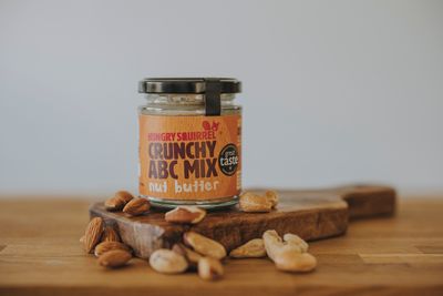 Jar of Crunchy ABC Mix nut butter on a wooden board with scattered almonds, brazils, and cashew nuts – natural ingredients