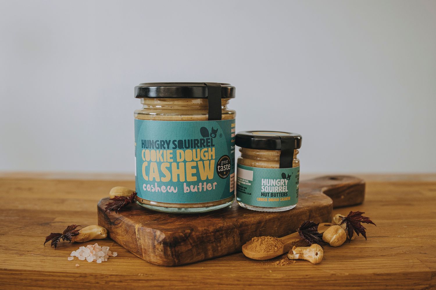 Cookie Dough Cashew Butter 180g and 40g jars displayed on a wooden board with whole cashews.

