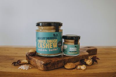 Cookie Dough Cashew Butter 180g and 40g jars displayed on a wooden board with whole cashews.

