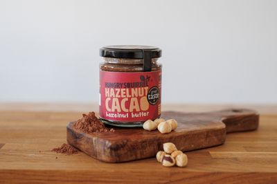 Jar of Hungry Squirrel Hazelnut Cacao butter on a wooden board beside whole hazelnuts.