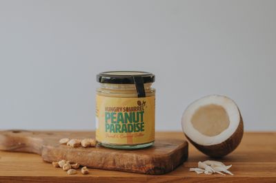 180g Peanut Paradise Nut Butter jar with yellow label on a wooden board, surrounded by coconut pieces.