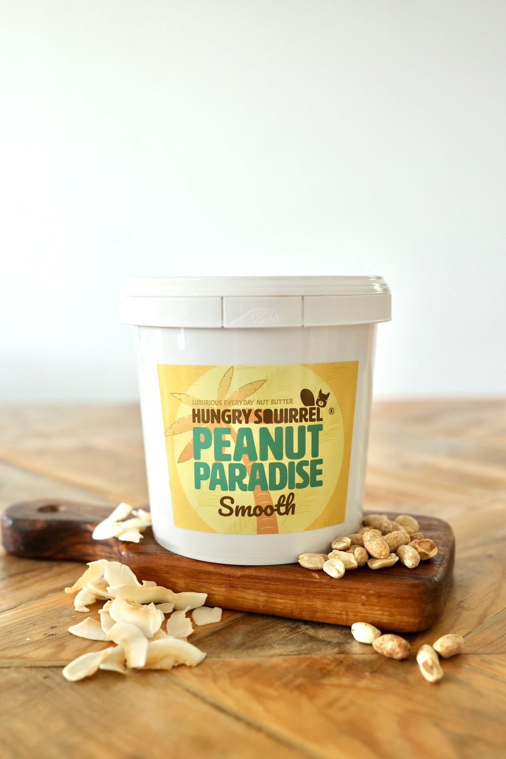 1kg Peanut Paradise Nut Butter tub with yellow label on a wooden surface, surrounded by peanuts.