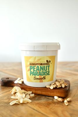 1kg Peanut Paradise Nut Butter tub with yellow label on a wooden surface, surrounded by peanuts.