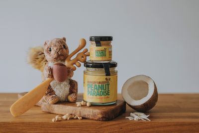 Peanut Paradise Nut Butter jars (180g and 40g) stacked on a wooden surface with peanuts and coconut pieces beside them.