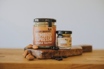 Almond Espresso Nut Butter 180g jar and 40g taster jar displayed on a wooden board with coffee beans and almonds.