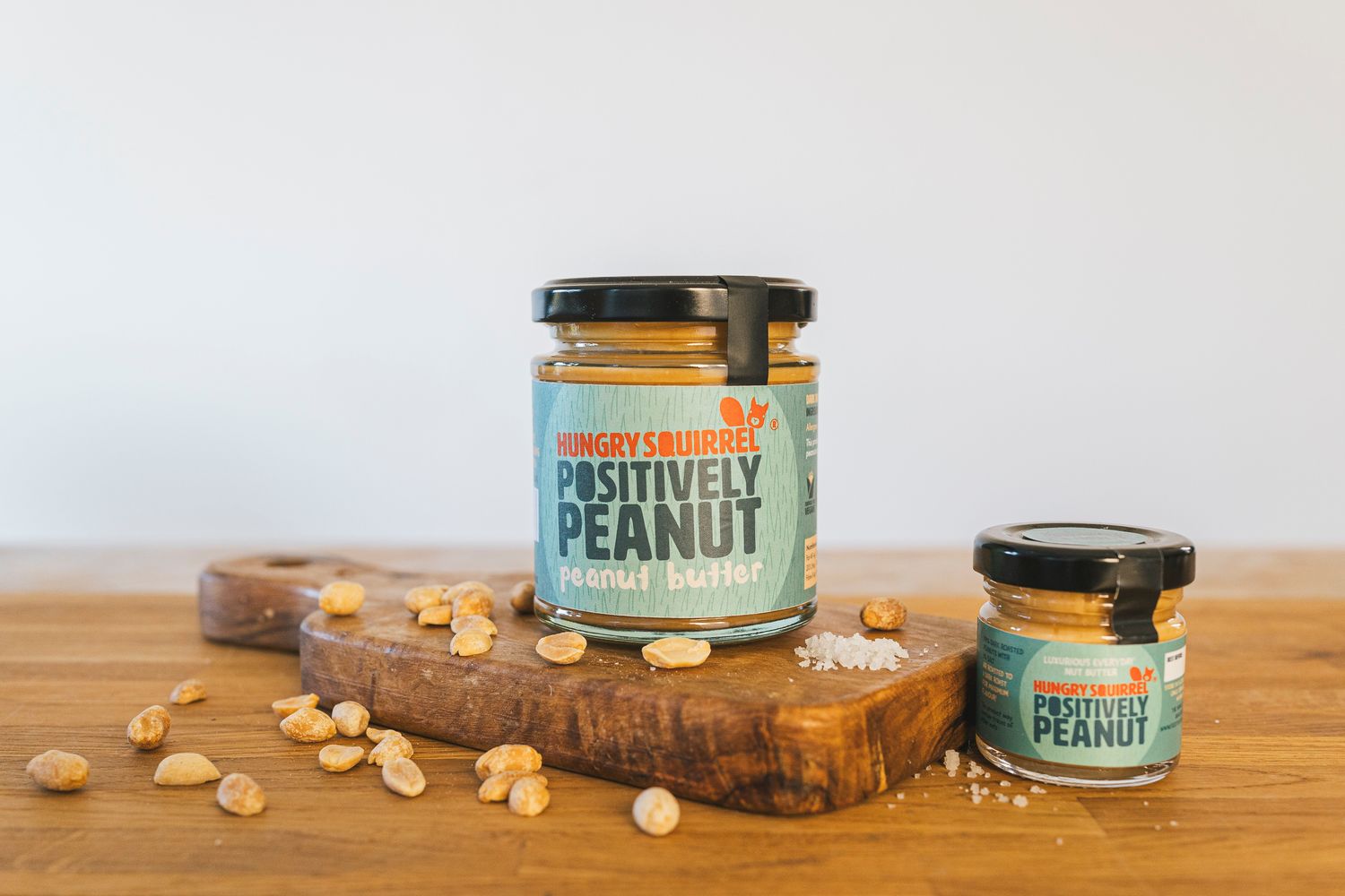 180g jar of Positively Peanut Butter with teal label on a wooden board, surrounded by peanuts.