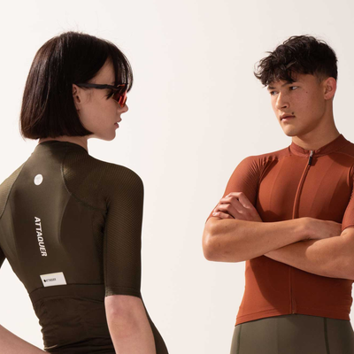 Two cyclists stand facing each other wearing Attaquer cycling jersey to display the front and back designs. The girl wears cycling glasses.